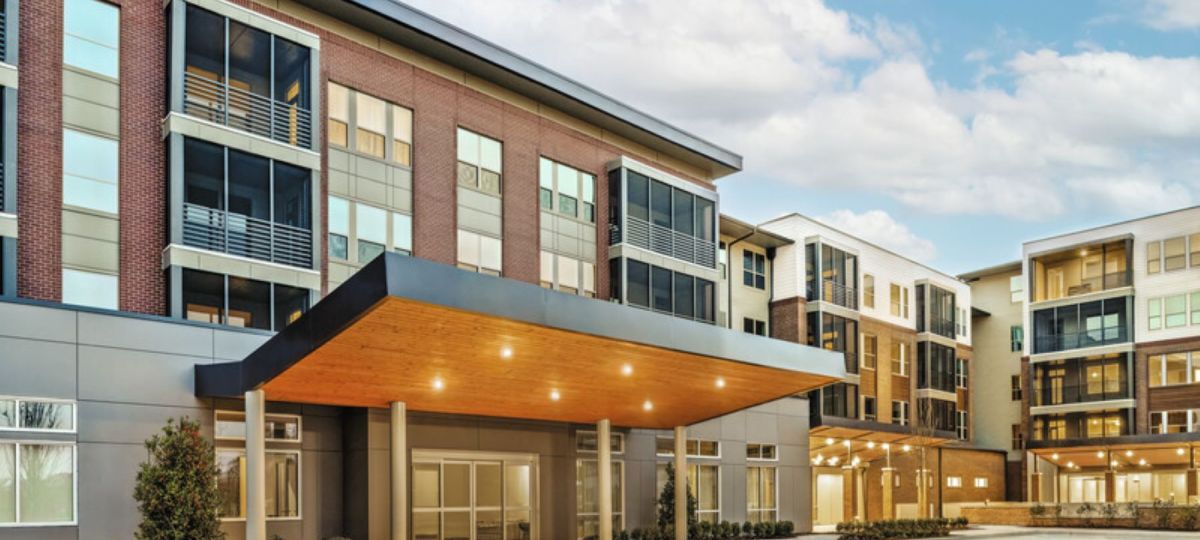 Modern senior living community with covered entryway, large windows, and multi-story brick and panel exterior under a blue sky.