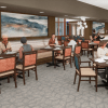 Spacious senior living dining room with multiple tables, cushioned chairs, wall art, and residents enjoying a meal.